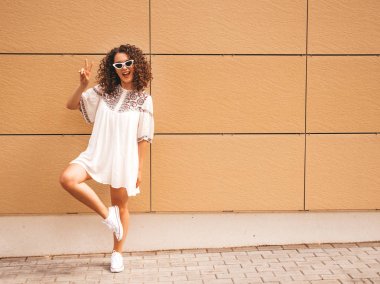 Yaz hipster beyaz elbise giymiş afro bukleler saç modeli ile güzel gülümseyen model. Seksi kaygısız kız güneş gözlüğü sarı duvar a yakın sokakta poz. Pozitif kadın eğleniyor. Barış işaretini gösterir