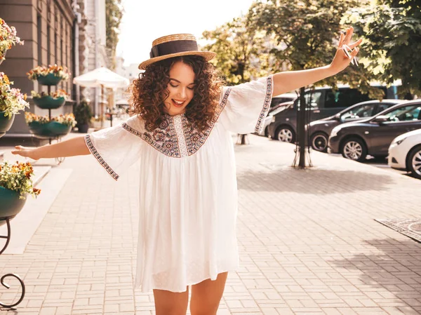 Beautiful smiling model with afro curls hairstyle dressed in summer hipster white dress. Sexy carefree girl posing in the street background. Trendy funny and positive woman having fun.Raising hands