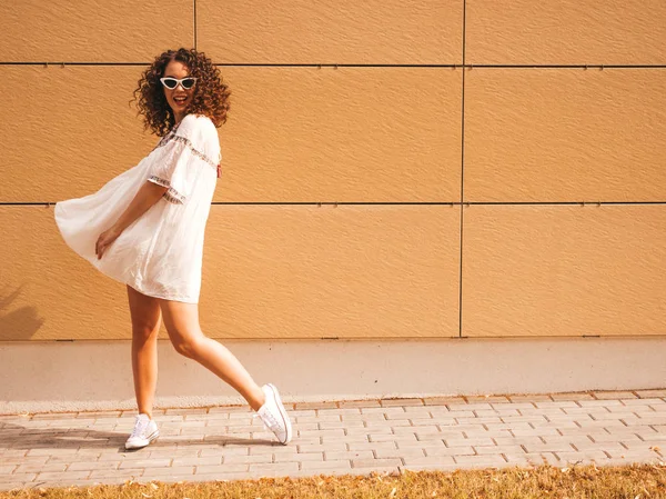 Beautiful smiling model with afro curls hairstyle dressed in summer hipster white dress.Sexy carefree girl posing in street near yellow wall in sunglasses.Funny and positive woman having fun