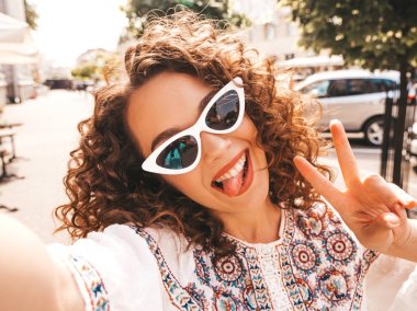 Yaz hipster beyaz elbise giymiş afro bukleler saç modeli ile güzel gülümseyen model. Seksi kaygısız kız güneş gözlüğü sokakta poz. Akıllı telefonda selfie selfie portre fotoğrafları çekmek