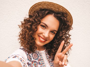Yaz hipster beyaz elbise giymiş afro bukleler saç modeli ile güzel gülümseyen model. Seksi kaygısız kız whtie duvara yakın sokakta poz. Şapkalı akıllı telefonda selfie selfie portre fotoğrafları çekmek