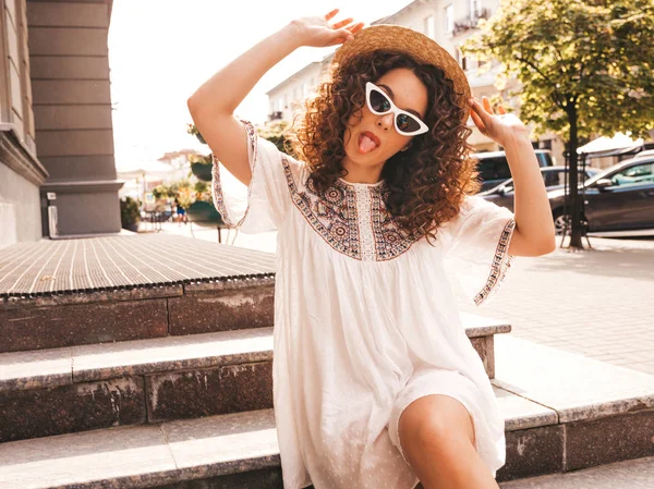Beautiful smiling model with afro curls hairstyle dressed in summer hipster white dress.Sexy carefree girl posing in street.Trendy and positive woman having fun in sunglasses.Shows peace sign in hat