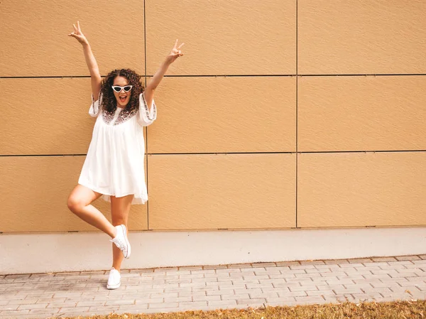 Beautiful smiling model with afro curls hairstyle dressed in summer hipster white dress.Sexy carefree girl posing in street near yellow wall in sunglasses.Positive woman having fun.Raising hands