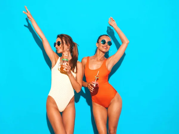 夏季五颜六色的泳装泳装中 两个美丽性感微笑的嬉皮士女人 戴着太阳镜的时髦女孩 发疯了在蓝色上孤立的有趣模型 喝新鲜的鸡尾酒举手 — 图库照片