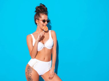 Hortlak saç modeli ile genç güzel seksi kadın. Güneş gözlüğü rahat yaz beyaz mayo Trendy kız. Sıcak model mavi izole. Yeme, ısırma şeker lolipop