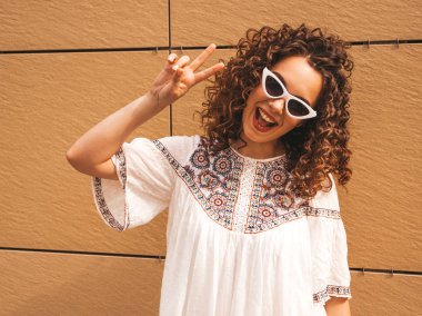 Yaz hipster beyaz elbise ve güneş gözlüğü giymiş afro bukleler saç modeli ile güzel gülümseyen model. Seksi kaygısız kız sarı duvara yakın sokakta poz. Komik kadın barış işareti gösterir