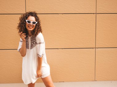 Yaz hipster beyaz elbise ve güneş gözlüğü giymiş afro bukleler saç modeli ile güzel gülümseyen model. Seksi kaygısız kız sarı duvara yakın sokakta poz. Komik ve pozitif kadın eğleniyor
