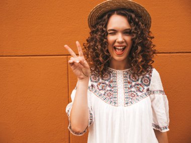 Yaz hipster beyaz elbise ve şapka giymiş afro bukleler saç modeli ile güzel gülümseyen model. Seksi kaygısız kız sarı duvara yakın sokakta poz. olumlu kadın eğlenmek, barış işareti gösterir