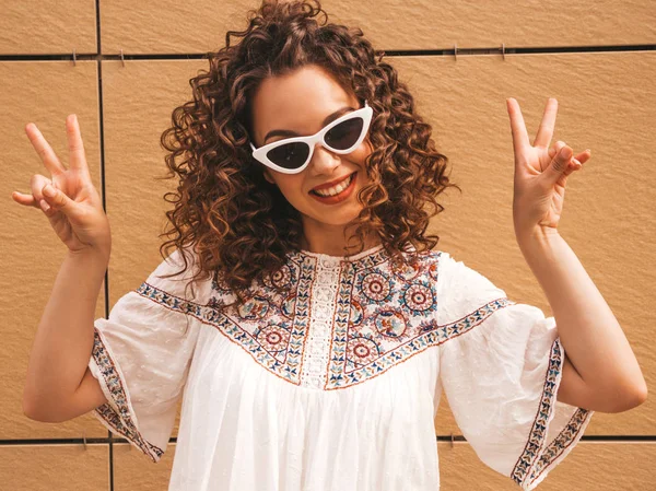 Beautiful smiling model with afro curls hairstyle dressed in summer hipster white dress and sunglasses.Sexy carefree girl posing in the street near yellow wall.Funny woman shows peace sign