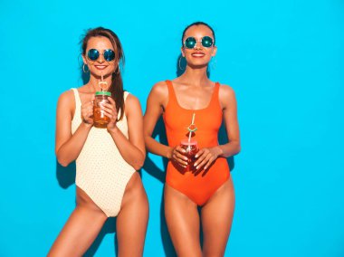 Yaz renkli mayo mayo iki güzel seksi gülümseyen hipster kadın. Güneş gözlükleri trendy kızlar. Deliriyorum. Mavi üzerine izole edilmiş komik modeller. İçme taze kokteyl smoozy içecek