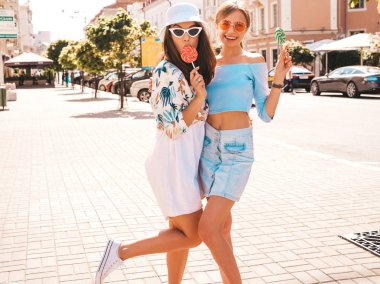 Trendy yazlık giysiler ve panama şapka iki genç güzel gülümseyen hipster kız. Seksi kaygısız kadınlar sokak arka planda poz. Güneş gözlüğü ile eğlenen pozitif modeller. Şeker lolipop yeme