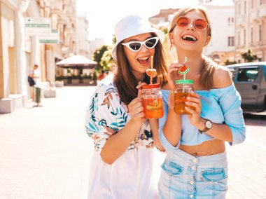 Trendy yazlık giysiler ve panama şapka iki genç güzel gülümseyen hipster kız. Sokakta poz veren seksi kaygısız kadınlar. Güneş gözlüğü ile eğlenen pozitif modeller. İçme taze kokteyl smoozy içecek