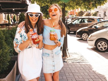 Trendy yazlık giysiler ve panama şapka iki genç güzel gülümseyen hipster kız. Sokakta poz veren seksi kaygısız kadınlar. Güneş gözlüğü ile eğlenen pozitif modeller. İçme taze kokteyl smoozy içecek