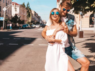 Gülümseyen güzel kız ve yakışıklı erkek arkadaşı. Gündelik yazlık elbiseli kadın ve kot elbiseli adam. Mutlu, neşeli bir aile. Kadın arka planda eğleniyor. Güneş gözlüklü çifte sarılıyor.