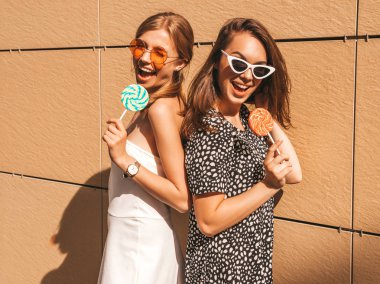 Trendy yaz elbisesi iki genç güzel gülümseyen hipster kız. Seksi kaygısız kadınlar sarı duvarın yakınındaki sokakta poz. Güneş gözlüğü ile eğlenen pozitif modeller. Şeker lolipop yeme