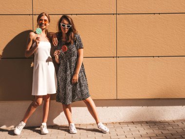 Trendy yaz elbisesi iki genç güzel gülümseyen hipster kız. Seksi kaygısız kadınlar sarı duvarın yakınındaki sokakta poz. Güneş gözlüğü ile eğlenen pozitif modeller. Şeker lolipop yeme