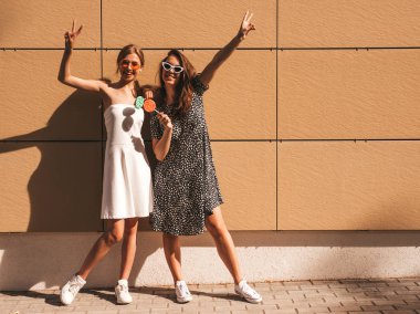 Trendy yaz elbisesi iki genç güzel gülümseyen hipster kız. Seksi kaygısız kadınlar sarı duvarın yakınındaki sokakta poz. Güneş gözlüğü ile eğlenen pozitif modeller. Şekerli lolipop yiyorum. El kaldırma