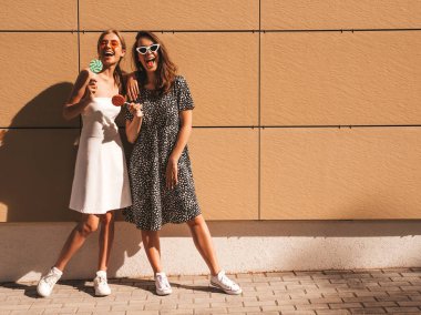 Trendy yaz elbisesi iki genç güzel gülümseyen hipster kız. Seksi kaygısız kadınlar sarı duvarın yakınındaki sokakta poz. Güneş gözlüğü ile eğlenen pozitif modeller. Şeker lolipop yeme