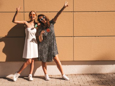 Trendy yaz elbisesi iki genç güzel gülümseyen hipster kız. Seksi kaygısız kadınlar sarı duvarın yakınındaki sokakta poz. Güneş gözlüğü ile eğlenen pozitif modeller. Şekerli lolipop yiyorum. El kaldırma