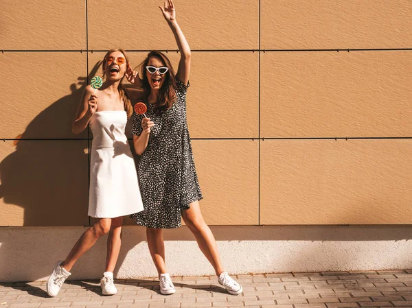 Trendy yaz elbisesi iki genç güzel gülümseyen hipster kız. Seksi kaygısız kadınlar sarı duvarın yakınındaki sokakta poz. Güneş gözlüğü ile eğlenen pozitif modeller. Şekerli lolipop yiyorum. El kaldırma