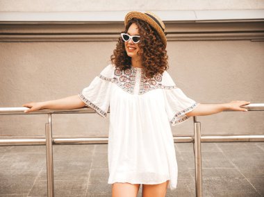 Yaz hipster beyaz elbise giymiş Afro bukleler saç ile güzel gülümseyen model. Seksi kaygısız kız sokakta poz. Şık komik ve pozitif kadın güneş gözlüğü ve şapka eğlenmek