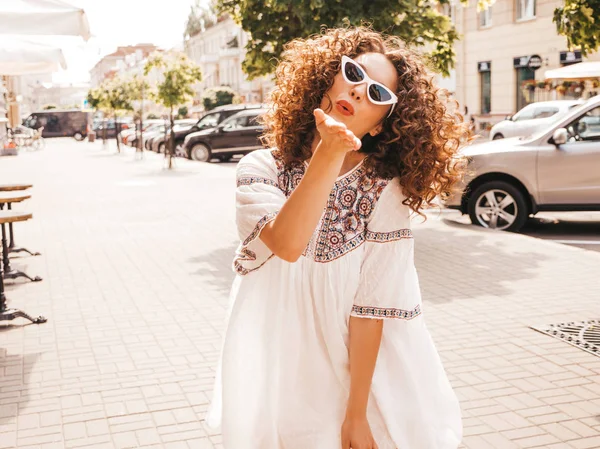 Beautiful smiling model with afro curls hairstyle dressed in summer hipster white dress.Sexy carefree girl posing in street.Trendy funny and positive woman having fun in sunglasses.Gives air kiss