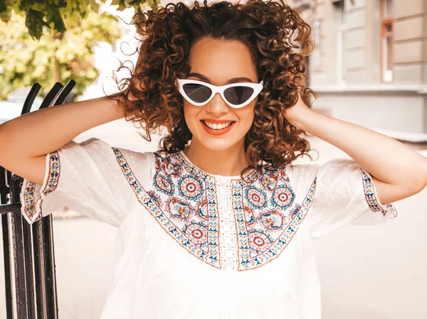 Beautiful smiling model with afro curls hairstyle dressed in summer hipster white dress.Sexy carefree girl posing in street.Trendy funny and positive woman having fun in sunglasses