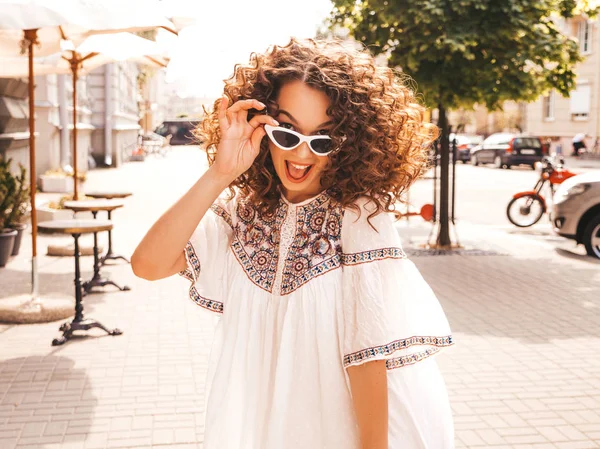 Beautiful smiling model with afro curls hairstyle dressed in summer hipster white dress.Sexy carefree girl posing in street.Trendy funny and positive woman having fun.Taking off sunglasses