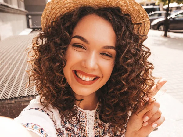 Beautiful smiling model with afro curls hairstyle dressed in summer hipster white dress.Sexy carefree girl posing in the street.Taking selfie self portrait photos in hat on smartphone
