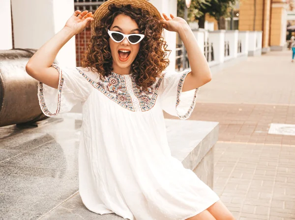 Beautiful smiling model with afro curls hairstyle dressed in summer hipster white dress.Sexy carefree girl posing in street.Trendy funny and positive woman having fun in sunglasses and hat