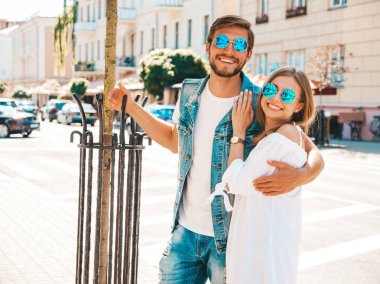 Gülümseyen güzel kız ve yakışıklı erkek arkadaşı. Gündelik yazlık elbiseli kadın ve kot elbiseli adam. Mutlu, neşeli bir aile. Kadın arka planda eğleniyor. Güneş gözlüklü çifte sarılıyor.