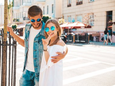 Gülümseyen güzel kız ve yakışıklı erkek arkadaşı. Gündelik yaz elbisesi giymiş bir kadın. Kot pantolon giymiş bir adam. Mutlu, neşeli bir aile. Kadın arka planda eğleniyor. Güneş gözlükleriyle barış işareti nasıl oluyor?