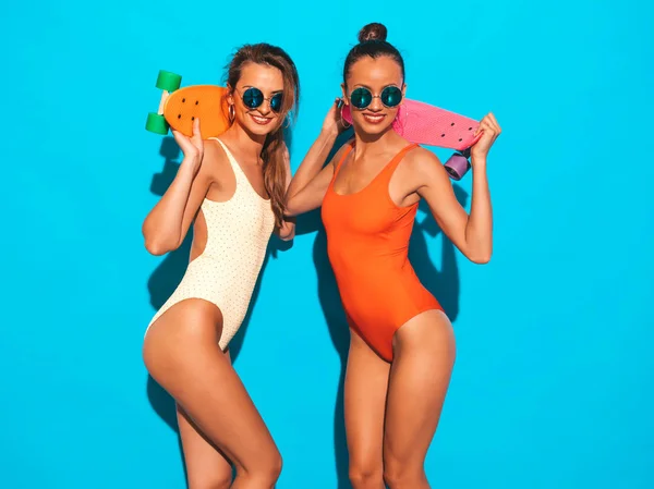 夏のカラフルな水着水着で2美しいセクシーな笑顔ヒップスターの女性 サングラスをかけたトレンディな女の子 カラフルなペニースケートボードを楽しむポジティブなモデル 青色で隔離 — ストック写真