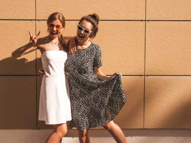Moda yazlık elbiseler iki genç güzel gülümseyen hipster kız. Seksi kaygısız kadınlar sarı duvarın yakınındaki sokakta poz. Güneş gözlüğü ile eğlenen pozitif modeller. Rock and roll işaretini gösterir