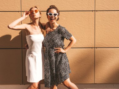 Moda yazlık elbiseler iki genç güzel gülümseyen hipster kız. Seksi kaygısız kadınlar sarı duvarın yakınındaki sokakta poz. Güneş gözlüğü ile eğlenen pozitif modeller