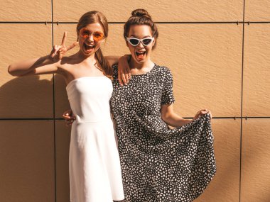Moda yazlık elbiseler iki genç güzel gülümseyen hipster kız. Seksi kaygısız kadınlar sarı duvarın yakınındaki sokakta poz. Güneş gözlüğü ile eğlenen pozitif modeller. Barış işaretini gösterir