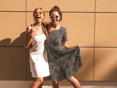 Moda yazlık elbiseler iki genç güzel gülümseyen hipster kız. Seksi kaygısız kadınlar sarı duvarın yakınındaki sokakta poz. Güneş gözlüğü ile eğlenen pozitif modeller. Rock and roll işaretini gösterir