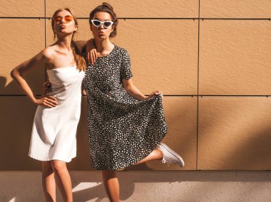 Moda yazlık elbiseler iki genç güzel gülümseyen hipster kız. Seksi kaygısız kadınlar sarı duvarın yakınındaki sokakta poz. Güneş gözlüğü ile eğlenen pozitif modeller. Ördek surat yapar