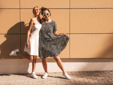Moda yazlık elbiseler iki genç güzel gülümseyen hipster kız. Seksi kaygısız kadınlar sarı duvarın yakınındaki sokakta poz. Güneş gözlüğü ile eğlenen pozitif modeller