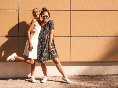 Moda yazlık elbiseler iki genç güzel gülümseyen hipster kız. Seksi kaygısız kadınlar sarı duvarın yakınındaki sokakta poz. Güneş gözlüğü ile eğlenen pozitif modeller