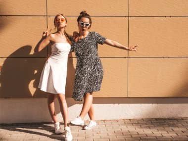 Moda yazlık elbiseler iki genç güzel gülümseyen hipster kız. Seksi kaygısız kadınlar sarı duvarın yakınındaki sokakta poz. Güneş gözlüğü ile eğlenen pozitif modeller. Barış işaretini gösterir