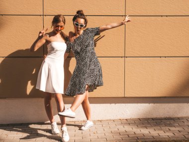 Moda yazlık elbiseler iki genç güzel gülümseyen hipster kız. Seksi kaygısız kadınlar sarı duvarın yakınındaki sokakta poz. Güneş gözlüğü ile eğlenen pozitif modeller. Barış işaretini gösterir