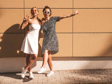 Moda yazlık elbiseler iki genç güzel gülümseyen hipster kız. Seksi kaygısız kadınlar sarı duvarın yakınındaki sokakta poz. Güneş gözlüğü ile eğlenen pozitif modeller. Barış işaretini gösterir