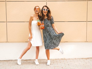 Moda yazlık elbiseler iki genç güzel gülümseyen hipster kız. Seksi kaygısız kadınlar sarı duvarın yakınındaki sokakta poz. Güneş gözlüğü yle eğlenen modeller. İçme taze kokteyl smoozy içecek