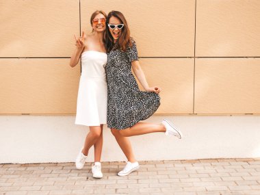 Moda yazlık elbiseler iki genç güzel gülümseyen hipster kız. Seksi kaygısız kadınlar sarı duvarın yakınındaki sokakta poz. Güneş gözlüğü ile eğlenen pozitif modeller. Barış işaretini gösterir