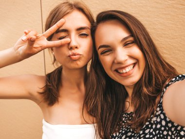 Yazlık kıyafetler giyen iki genç gülümseyen hipster kadın. Akıllı telefonda selfie selfie portre fotoğrafları çeken kızlar. Sarı duvarın yanındaki sokak arka planında poz veren mankenler. Kadın olumlu yüz duyguları gösteren