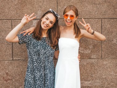 Moda yazlık elbiseler iki genç güzel gülümseyen hipster kız. Seksi kaygısız kadınlar sarı duvarın yakınındaki sokakta poz. Güneş gözlüğü ile eğlenen pozitif modeller. Barış işaretini gösterir