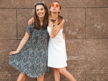 Moda yazlık elbiseler iki genç güzel gülümseyen hipster kız. Seksi kaygısız kadınlar kahverengi duvarın yakınındaki sokakta poz. Güneş gözlüğü ile eğlenen pozitif modeller. Hava öpücüğü vermek