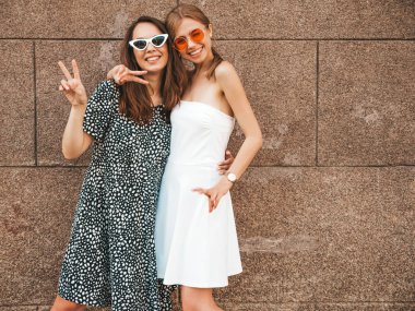 Moda yazlık elbiseler iki genç güzel gülümseyen hipster kız. Seksi kaygısız kadınlar sarı duvarın yakınındaki sokakta poz. Güneş gözlüğü ile eğlenen pozitif modeller. Barış işaretini gösterir