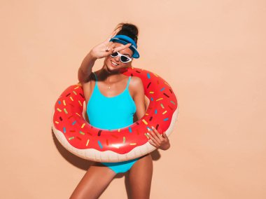 Young beautiful sexy smiling hipster woman in sunglasses.Girl in summer swimwear bathing suit with donut lilo inflatable mattress.Positive female going crazy.Near beige wall in transparent visor cap clipart
